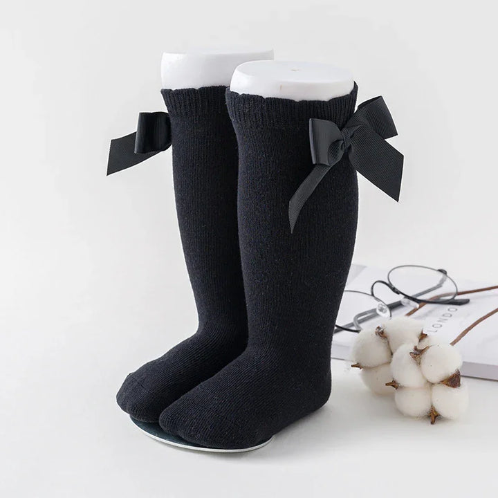 Black knee-high socks with decorative black bows on white mannequin feet, cotton branch and glasses nearby