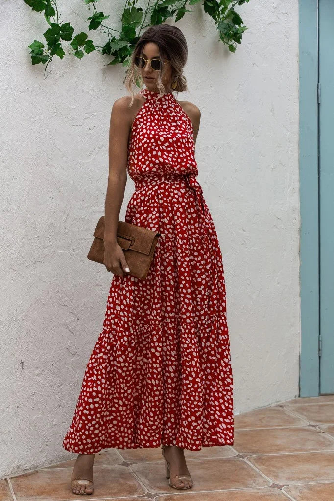 Woman in red polka dot halter maxi dress holding brown clutch on tiled patio with white wall and green plants