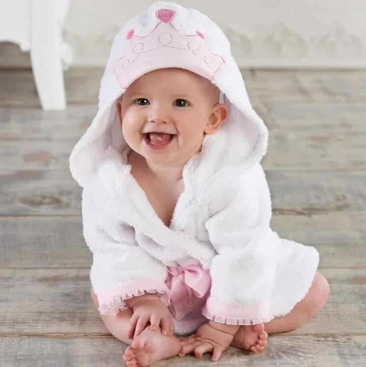 Smiling baby wearing white hooded bathrobe with pink crown and bow sitting on wooden floor