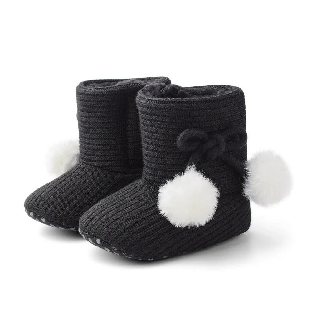 Black knitted baby booties with white pom-poms and non-slip soles on white background