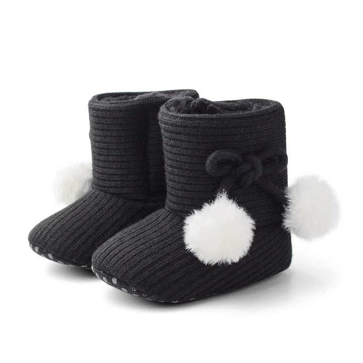 Black knitted baby booties with white pom-poms and non-slip soles on white background