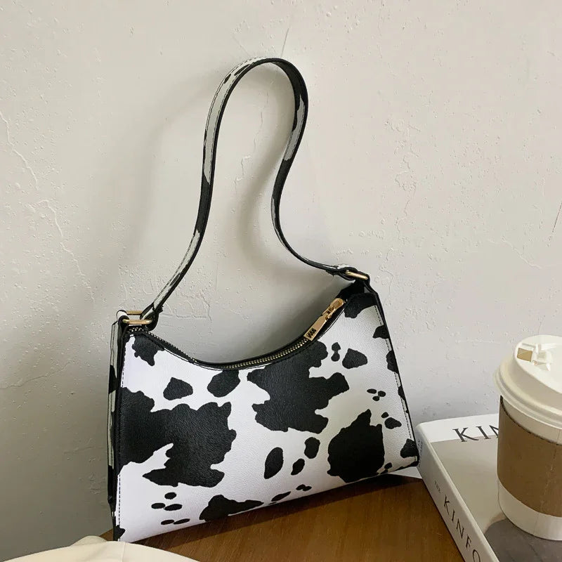 Black and white cow print shoulder bag with gold zipper on wooden table next to coffee cup and magazine