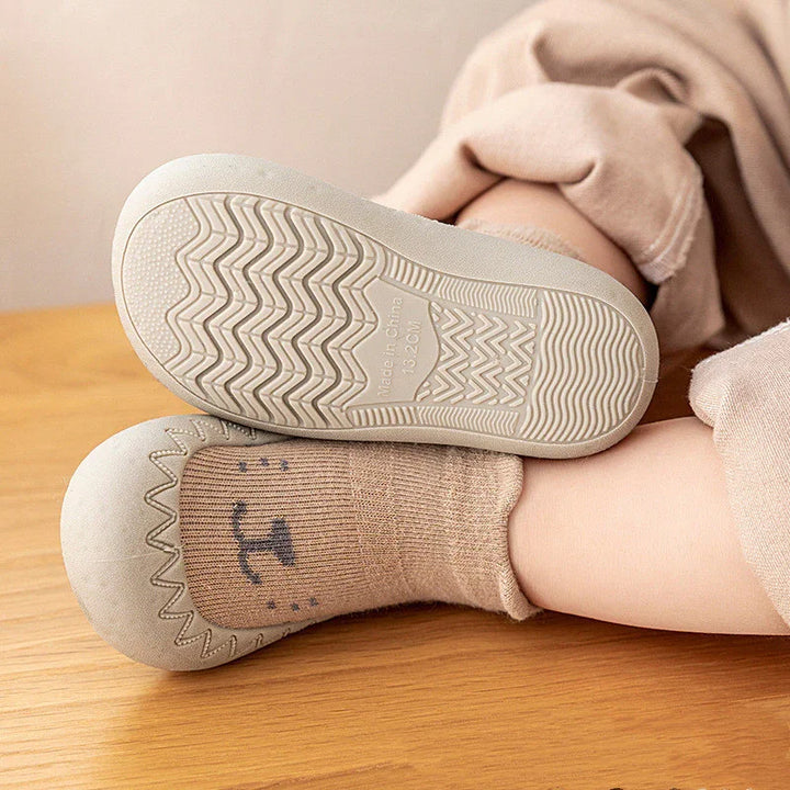 Baby wearing beige soft sole shoes with bear face socks on wooden floor