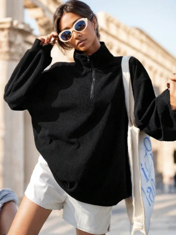 Young woman wearing oversized black quarter-zip sweater and white shorts with sunglasses in outdoor urban setting