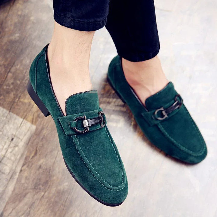 Men's green suede loafers with metal buckle detail worn with black pants on wooden floor