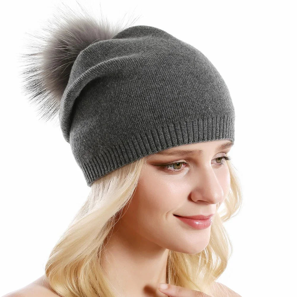 Blonde woman wearing a gray knit beanie with a fluffy pom pom against a white background
