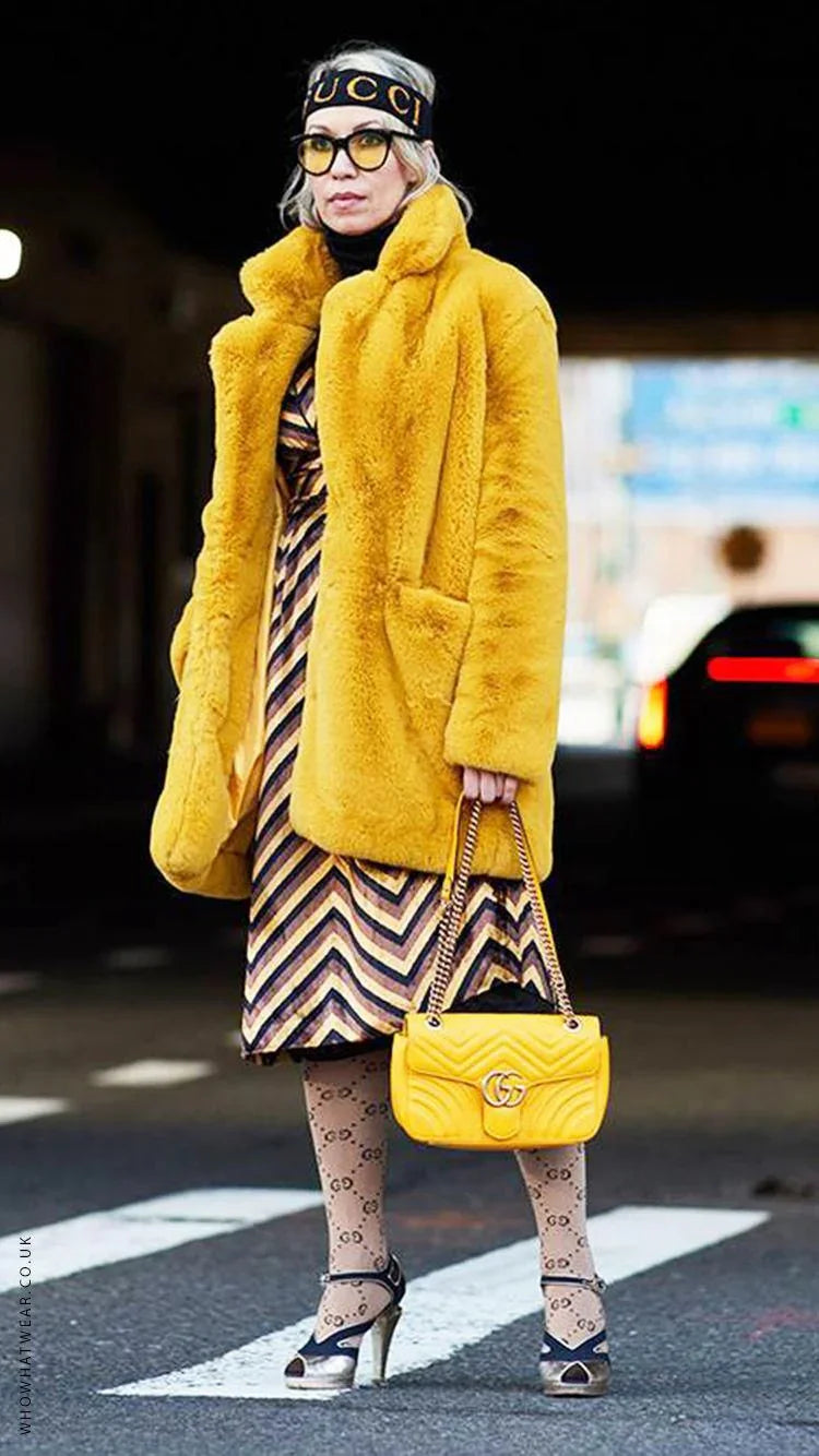 Woman wearing bright yellow faux fur coat, patterned dress, Gucci tights, and holding yellow Gucci handbag on street
