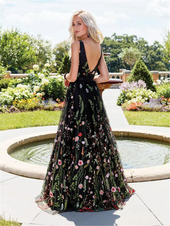 Blonde woman in black floral embroidered gown with deep V-back standing by garden fountain