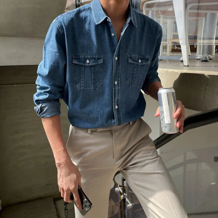 man wearing casual blue denim shirt and beige trousers holding a can and a smartphone indoors