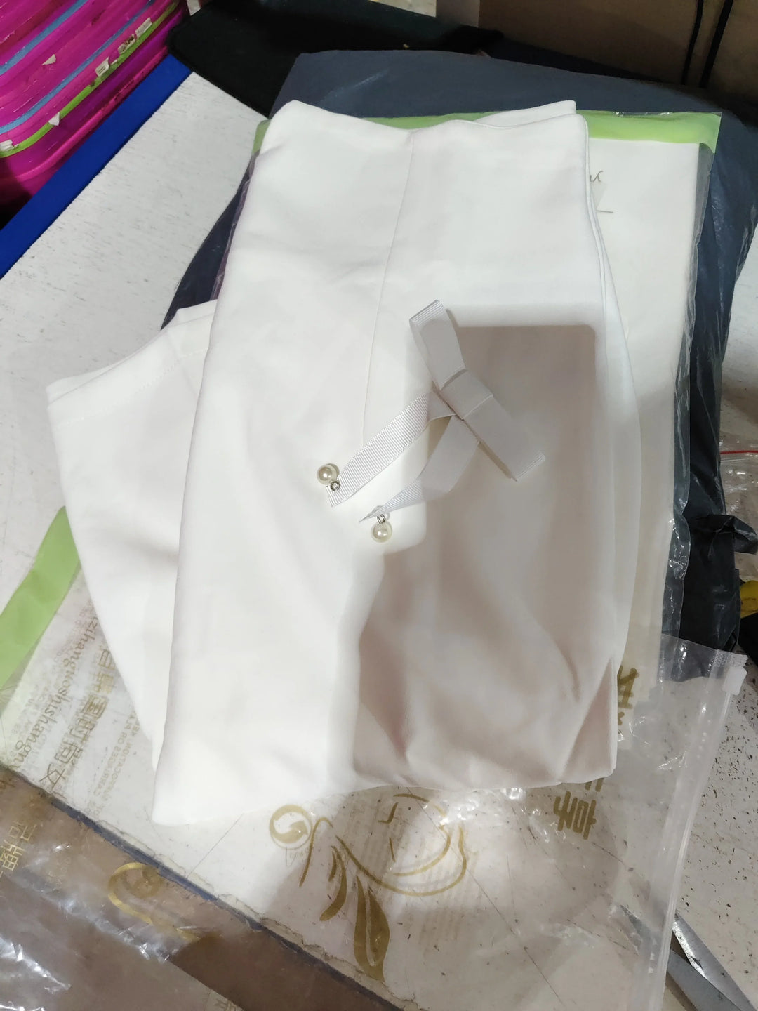 Folded white pants with a ribbon bow and pearl accents placed on packaging on a table
