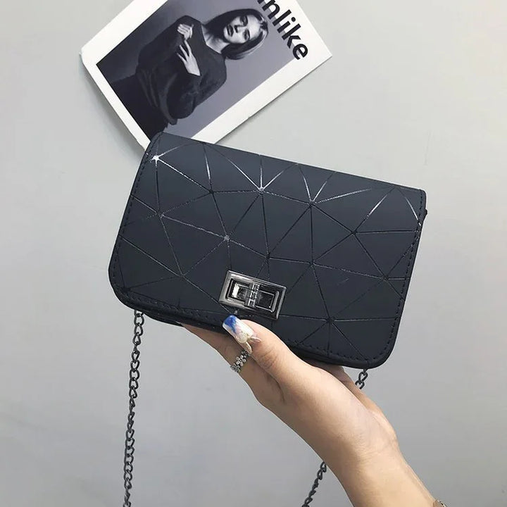 Hand holding black geometric pattern crossbody bag with silver chain strap against gray background and fashion magazine