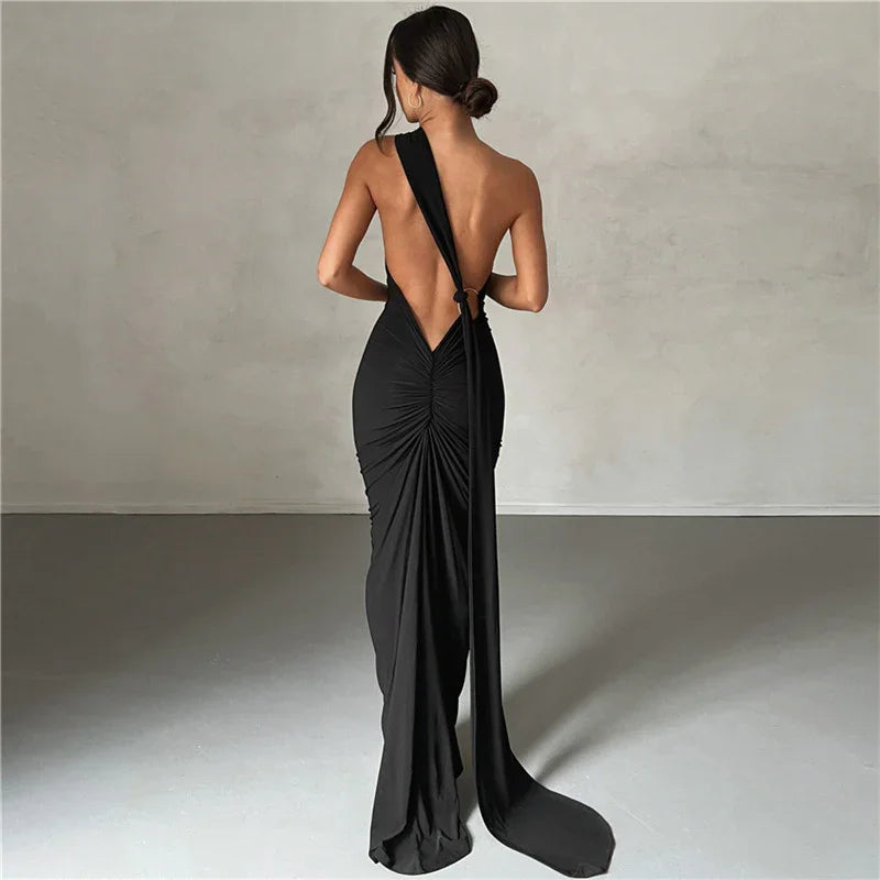 Woman in a sleek black backless gown with gathered fabric and long draped sash standing against gray wall