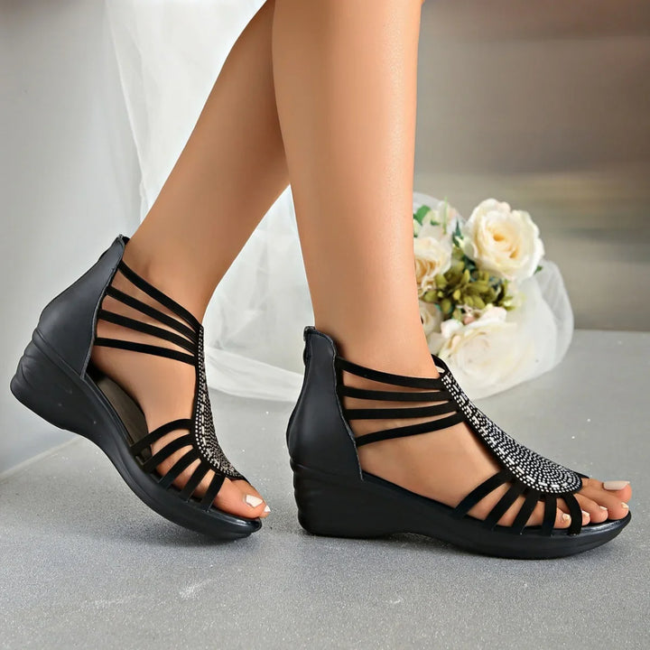 Black wedge sandals with strappy design and rhinestone embellishments on feet over grey floor