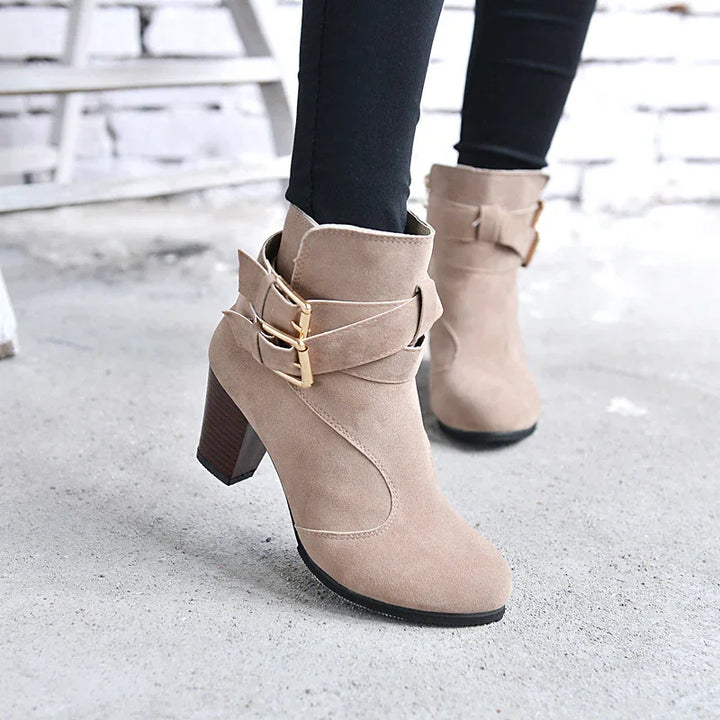 Beige suede ankle boots with gold buckles and block heels worn with black pants on concrete floor