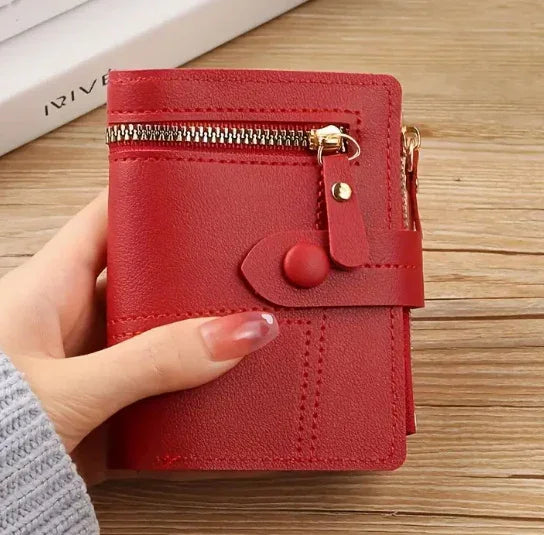 Hand holding red leather wallet with zipper pocket and snap button strap on wooden surface