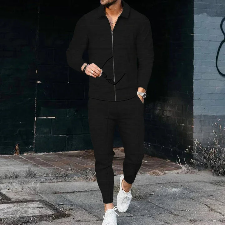 Man wearing a black zip-up jacket and jogger pants with white sneakers standing on an urban sidewalk