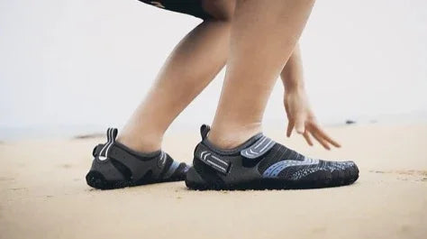 Person wearing black water shoes on sandy beach, barefoot-style footwear, beach activity