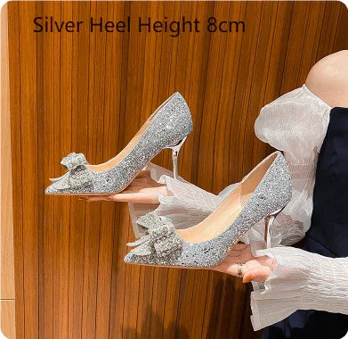 Woman holding silver glitter high heels with 8cm clear heels and decorative bow against wooden background