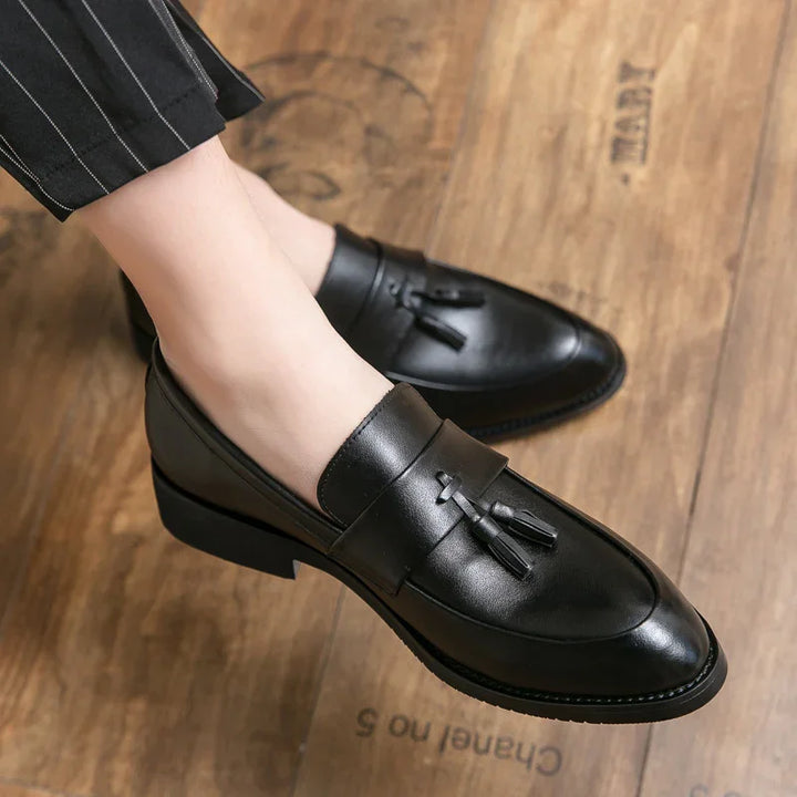 Black leather tassel loafers worn with pinstripe pants on wooden floor