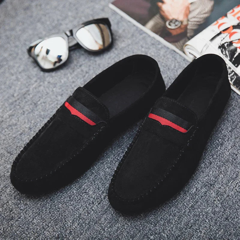 Black suede loafers with red and black stripe detail on grey carpet near reflective sunglasses and magazine