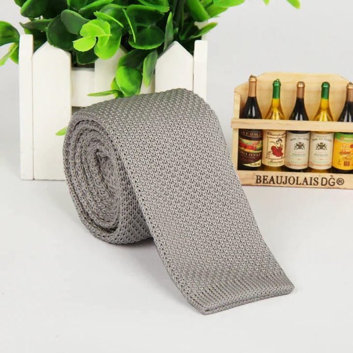 Rolled gray knitted tie on white surface with green plant and miniature wine bottle rack background