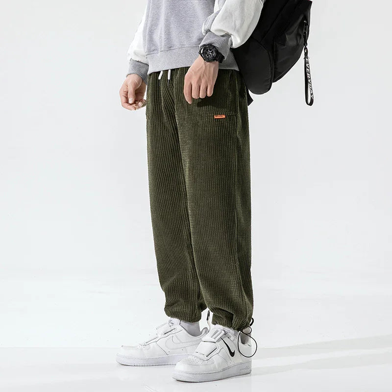 Man wearing olive green corduroy pants with grey sweatshirt and white Nike sneakers, carrying black backpack