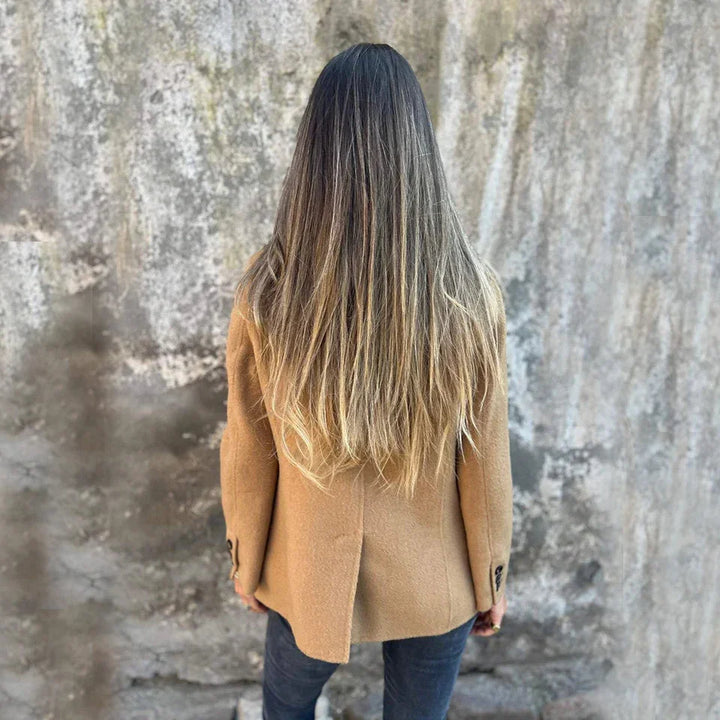 Woman with long ombre hair wearing camel coat and dark jeans standing against weathered concrete wall