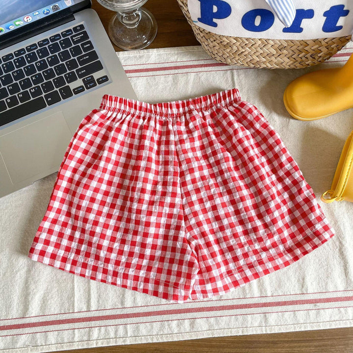 Red and white gingham checkered shorts on a cloth beside a laptop, basket, and yellow rain boots