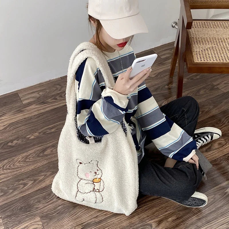 Person wearing striped long-sleeve shirt and cap, holding phone, with white fluffy bear tote bag sitting on wooden floor