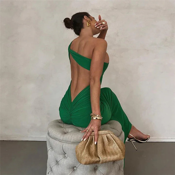 Woman in a backless green evening dress sitting on a tufted ottoman, holding a woven clutch handbag