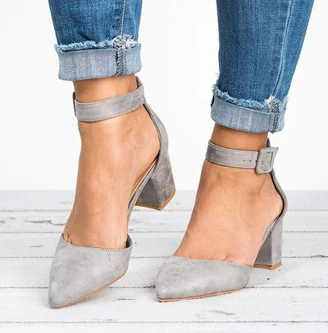 Grey suede block heel shoes with ankle straps worn with rolled-up blue jeans on white wooden floor