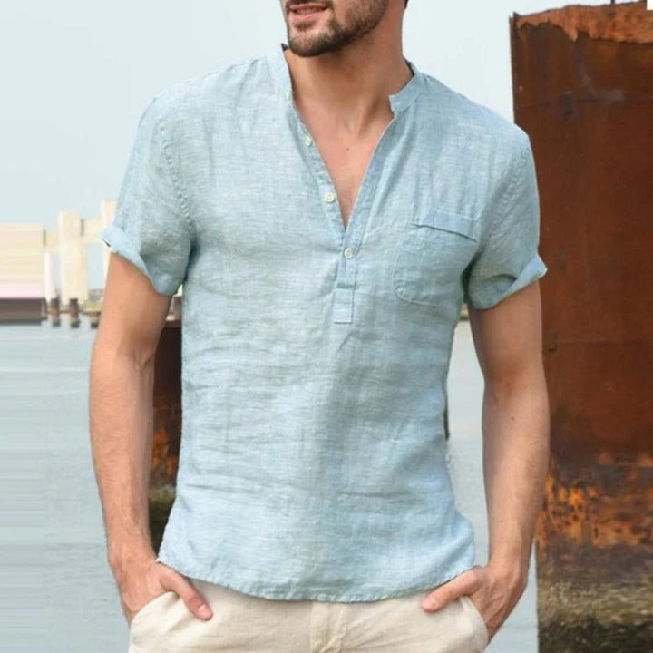 Man wearing light blue short-sleeve linen henley shirt with rolled sleeves near rusty metal and water