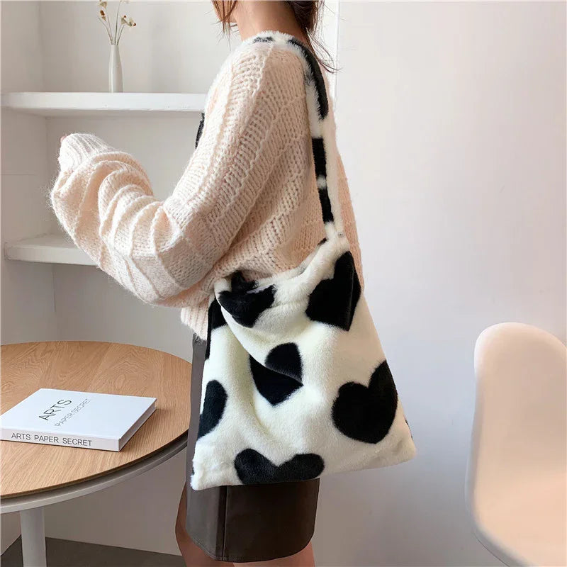 Person wearing beige knit sweater and black skirt holding black and white heart-patterned faux fur shoulder bag indoors
