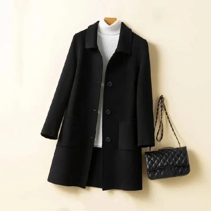 Black wool coat hanging with white turtleneck sweater and quilted black handbag on beige background