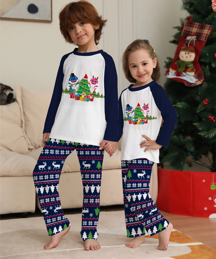 Two children wearing matching Christmas pajamas with holiday tree and reindeer pattern, standing by decorated Christmas tree