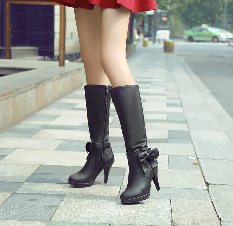 Woman wearing black high-heeled knee boots with bow detail on city sidewalk