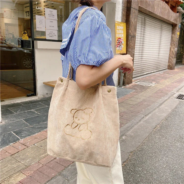 Woman in blue striped blouse holding beige corduroy tote bag with embroidered bear on city street