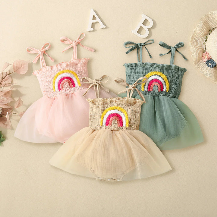 Three colorful baby girl dresses with rainbow embroidery and tulle skirts on beige background
