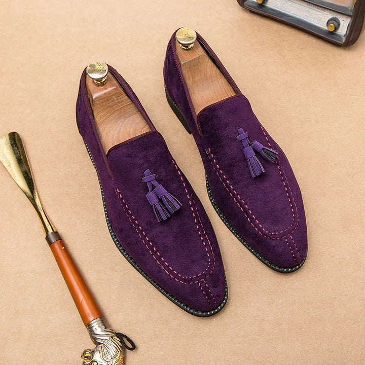 Purple suede tassel loafers with pink stitching and wooden shoe trees on beige surface