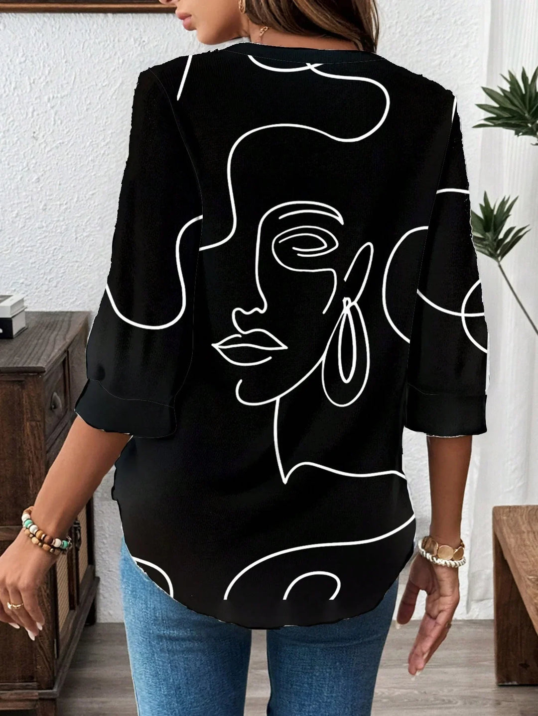 Woman wearing black blouse with white abstract face line art design, paired with blue jeans indoors