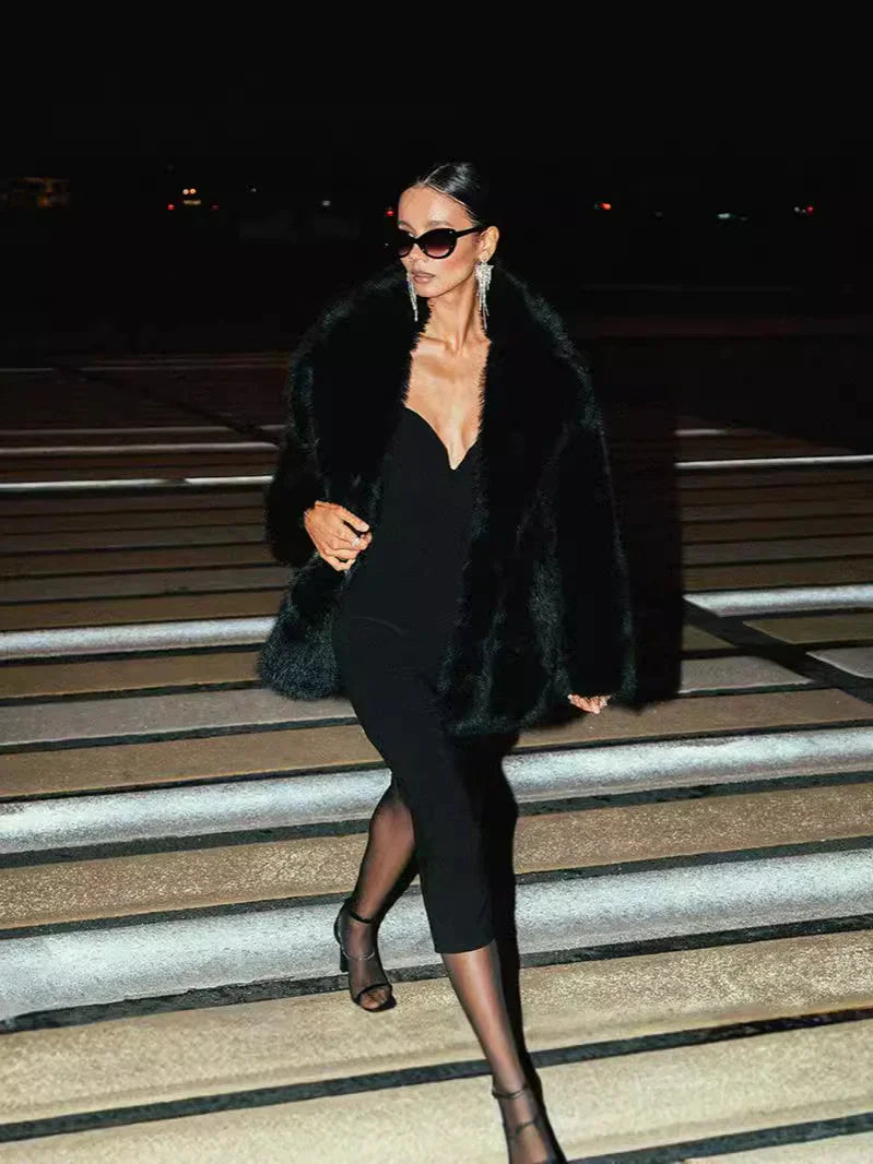Woman in black fur coat and dress walking on illuminated steps at night, wearing sunglasses and heels