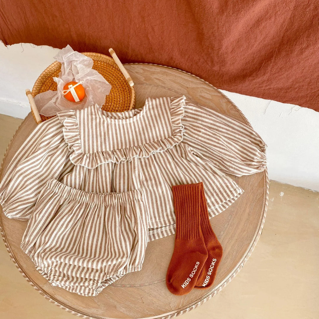 Striped beige baby outfit with ruffled top, matching bloomers, and rust-colored kids socks on wooden table