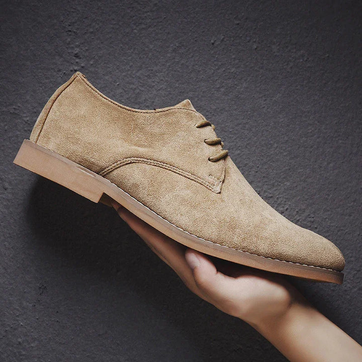 Hand holding a tan suede lace-up oxford shoe with a light brown sole against a gray textured background