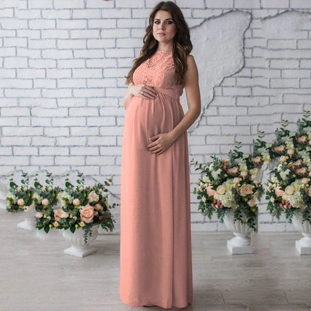 Pregnant woman in a peach lace maternity dress standing by floral arrangements in a white brick wall room