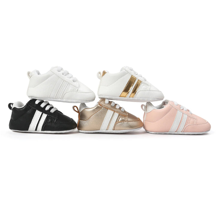 Five pairs of soft baby sneakers in white, black, metallic gold, and pink with stripes on a white background