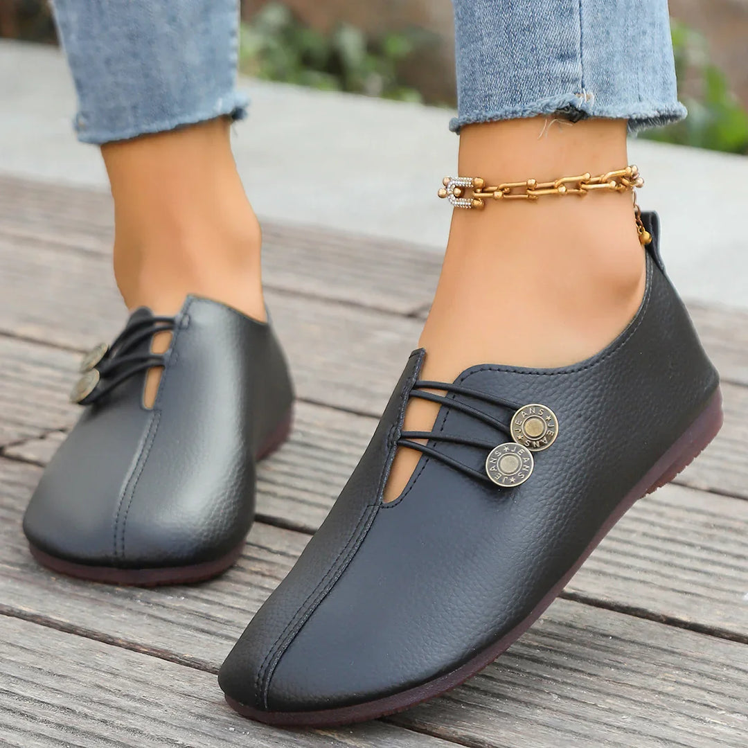 Black leather casual women's shoes with decorative buttons and ankle chain on wooden floor