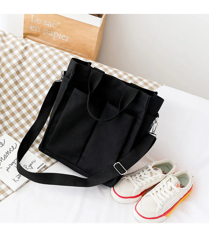 Black canvas tote bag with adjustable strap and front pockets on checkered fabric beside white sneakers with red and yellow stripes