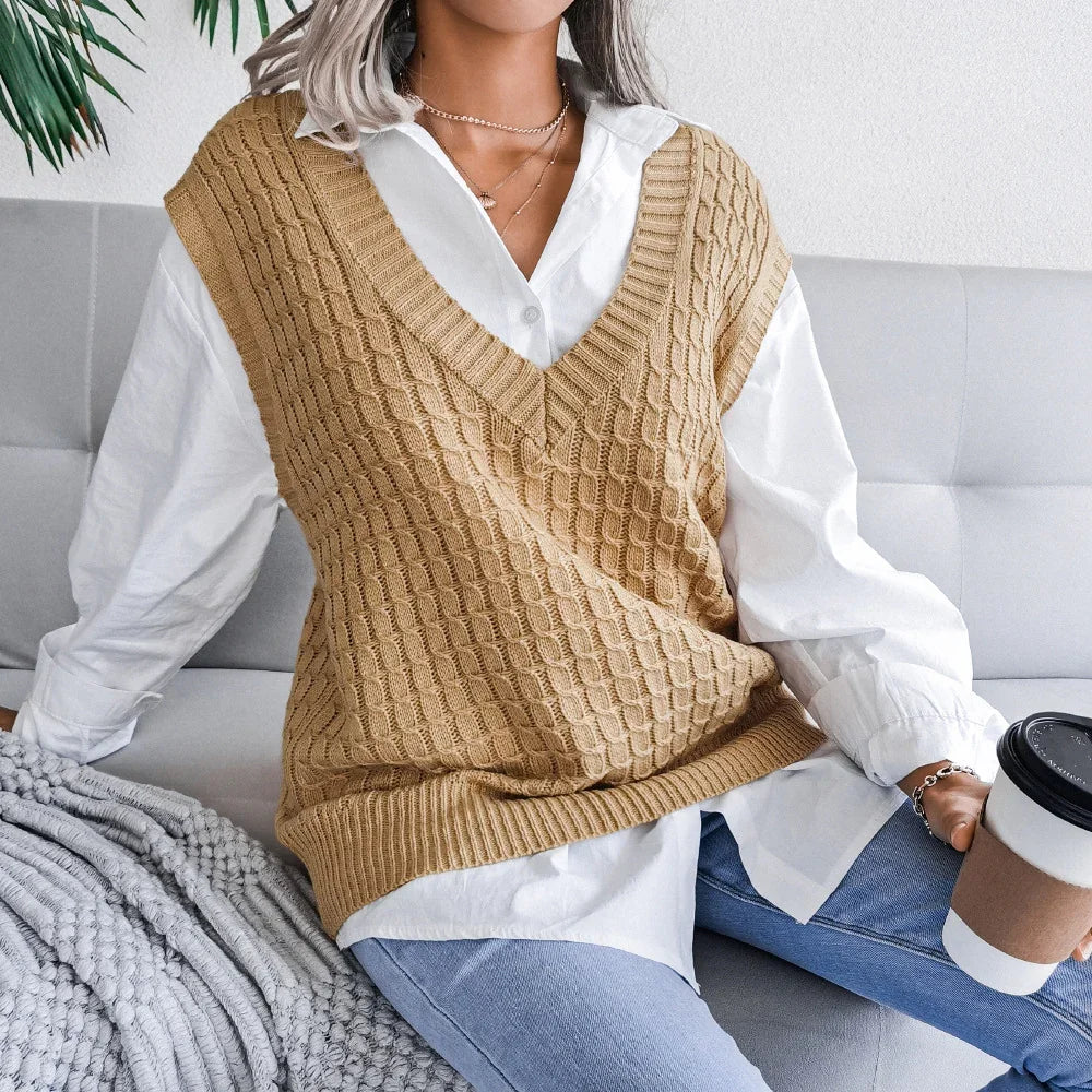 Woman sitting on gray sofa wearing tan knit vest over white shirt and blue jeans holding coffee cup