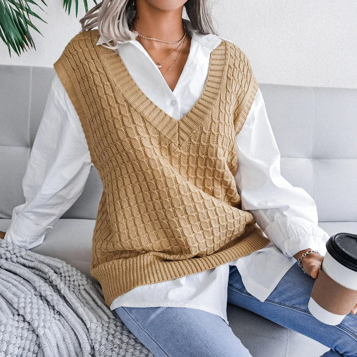 Woman sitting on gray sofa wearing tan knit vest over white shirt and blue jeans holding coffee cup