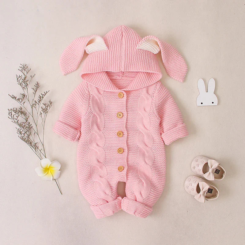 Pink knitted baby jumpsuit with bunny ears hood, surrounded by baby shoes, flower, and dried twigs